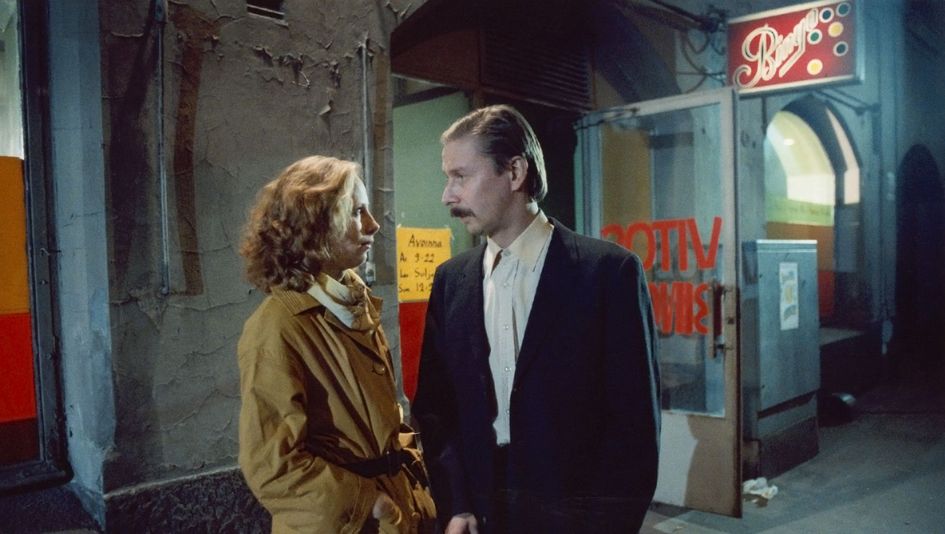 Film still from SHADOWS IN PARADISE: A man and a woman look at each other, behind them the illuminated display of a nightclub.