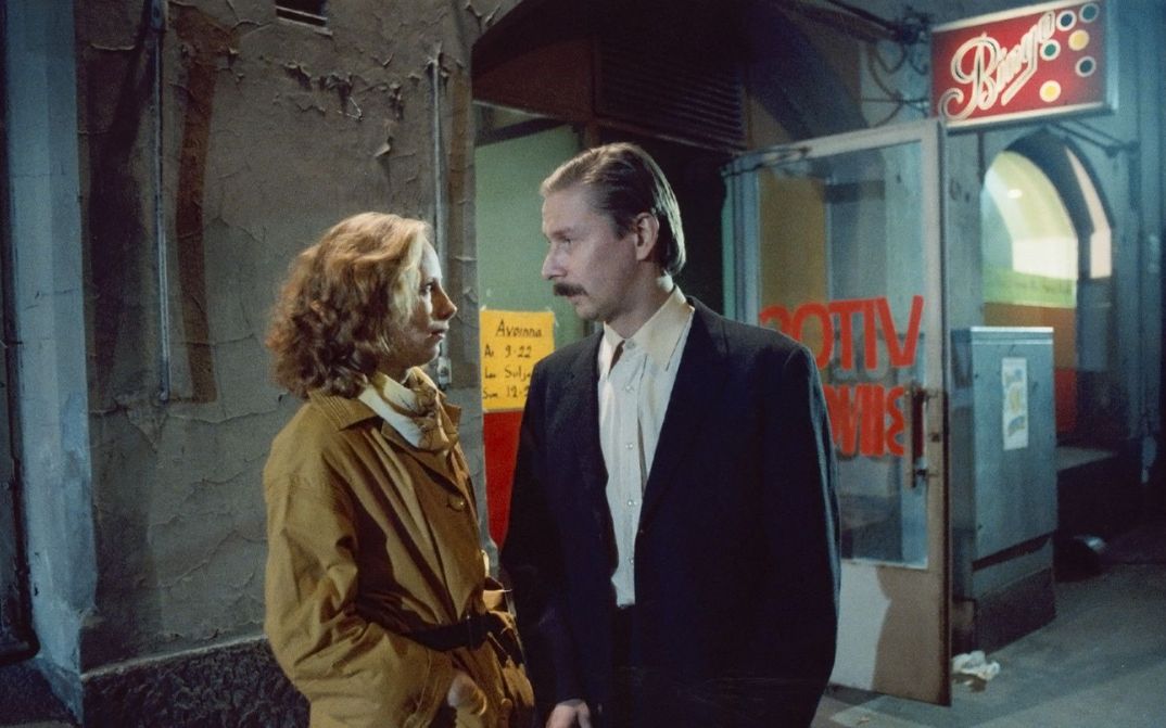 Filmstill aus SCHATTEN IM PARADIES: Ein Mann und eine Frau blicken sich an, hinter ihnen die beleuchtete Anzeige eines Nachtlokals.