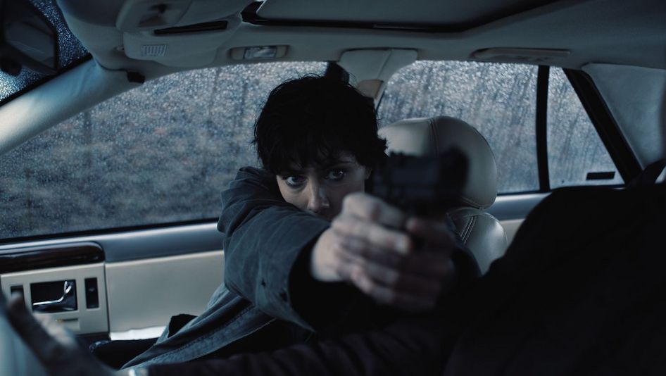 Filmstill aus LA MORT VIENDRA: Eine Person sitzt im Beifahrersitz eines Autos und hält ausgestreckt eine Waffe.