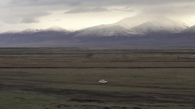 Film still from LANDSHAFT: A car drives through a landscape with snow-covered mountains in the background.