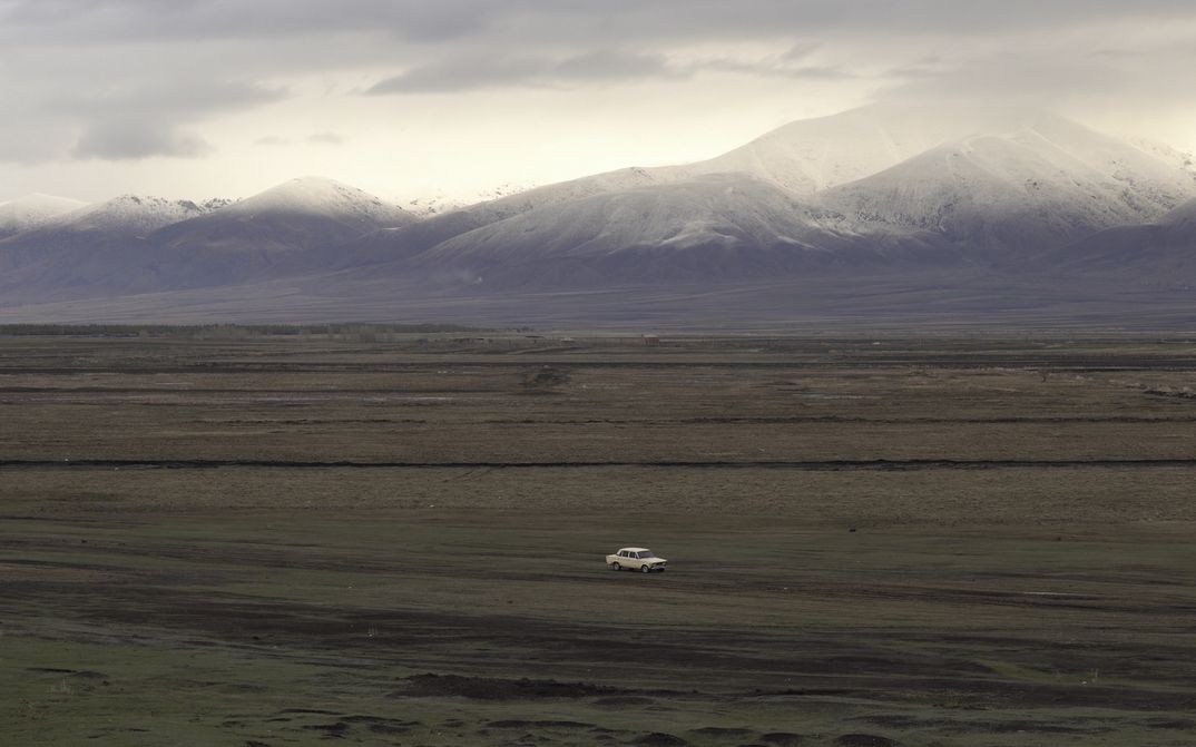 Film still from LANDSHAFT: A car drives through a landscape with snow-covered mountains in the background.