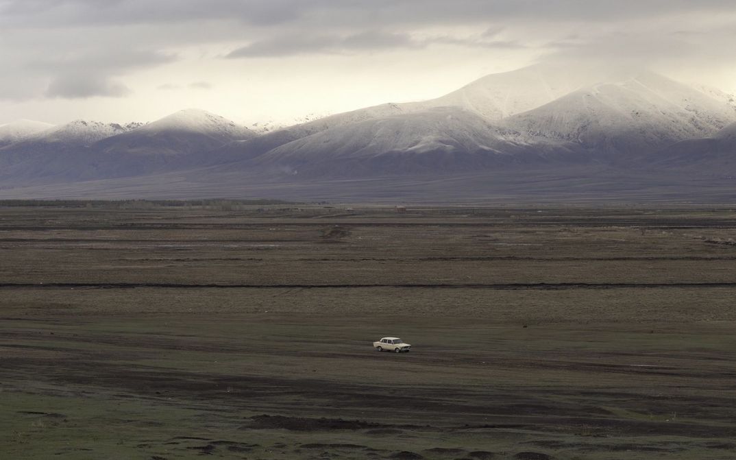 Filmstill aus LANDSHAFT: Ein Auto fährt durch eine karge Landschaft, im Hintergrund sind schneebedeckte Berge zu sehen.