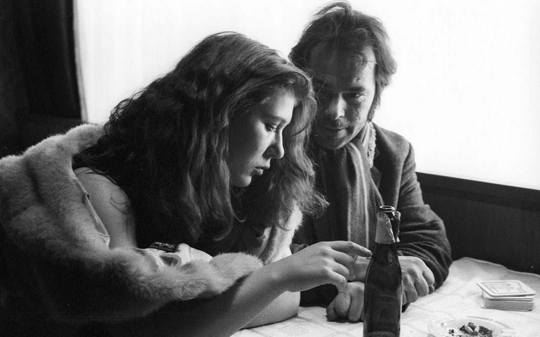 Film still from STROSZEK: A woman and a man are sitting at a table. She points at a beer bottle in front of her, he looks at the woman.