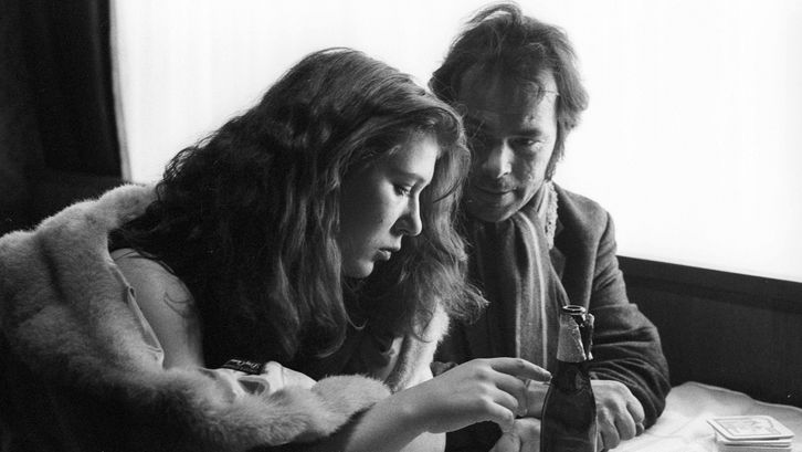 Film still from STROSZEK: A woman and a man are sitting at a table. She points at a beer bottle in front of her, he looks at the woman.