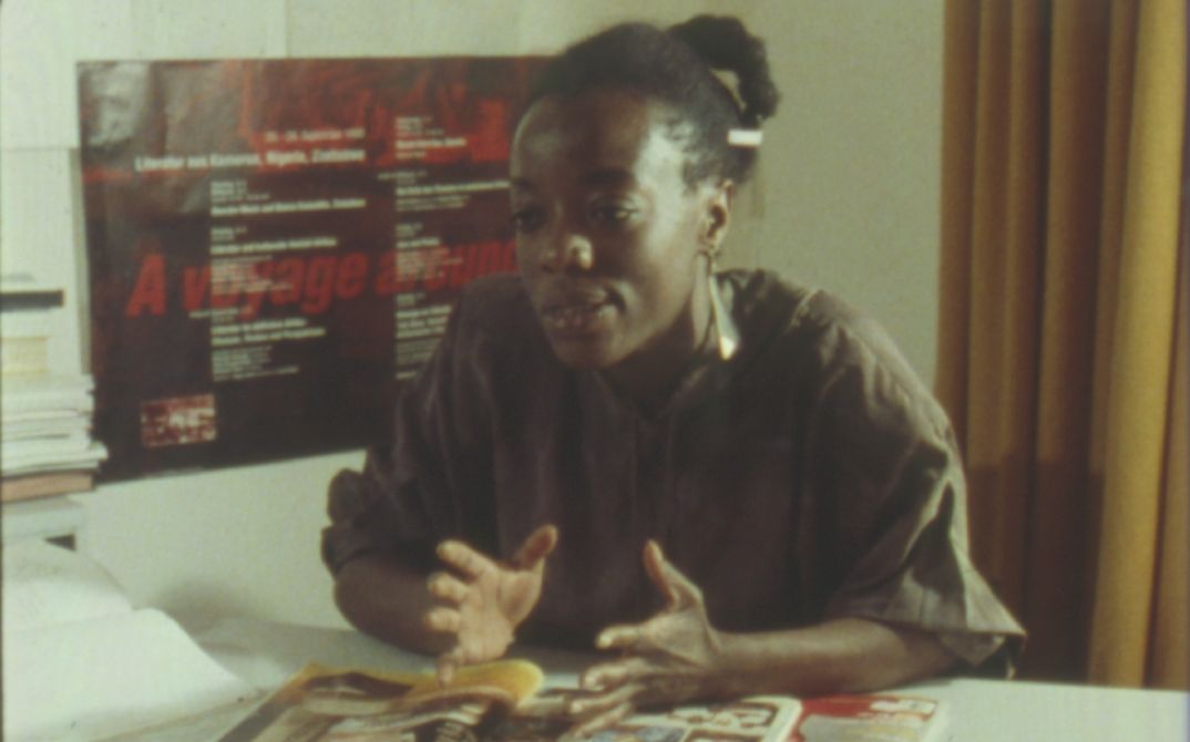 Film still from “Black in the Western World“. A woman is sitting at a desk, speaking to someone off camera. She gestures with her hands.