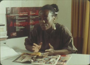 Film still from “Black in the Western World“. A woman is sitting at a desk, speaking to someone off camera. She gestures with her hands.