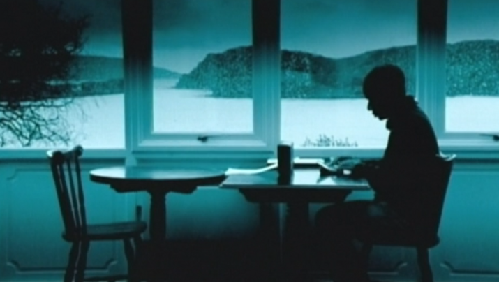 Filmstill aus DIGITOPIA: Man sieht das grünlich monochrom eingefärbte Bild eines Mannes, der am Tisch an einem großen Fenster sitzt. Hinter dem Fenster ist eine hügelige Landschaft zu sehen.