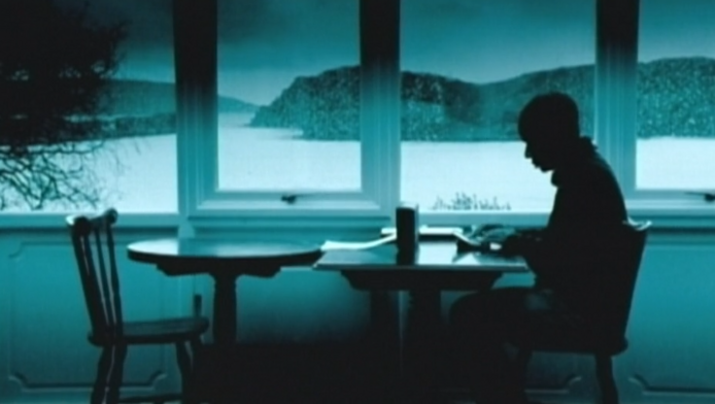 Filmstill aus DIGITOPIA: Man sieht das grünlich monochrom eingefärbte Bild eines Mannes, der am Tisch an einem großen Fenster sitzt. Hinter dem Fenster ist eine hügelige Landschaft zu sehen.