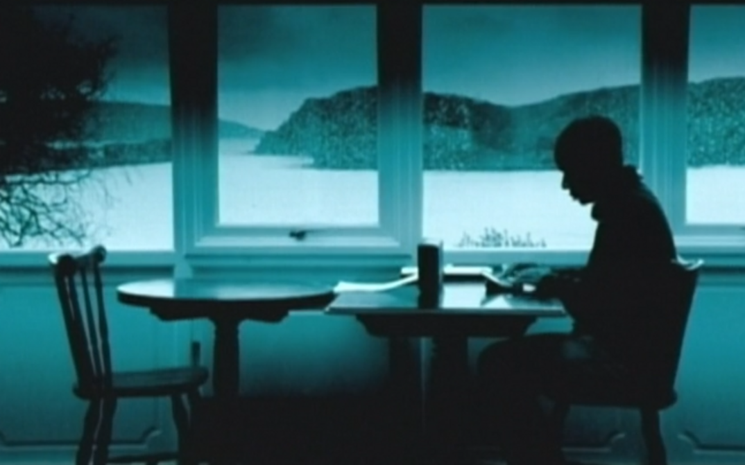 Film still from DIGITOPIA: You can see the greenish monochrome image of a man sitting at a table by a large window. A hilly landscape can be seen behind the window.