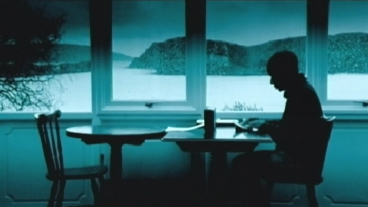 Film still from DIGITOPIA: You can see the greenish monochrome image of a man sitting at a table by a large window. A hilly landscape can be seen behind the window.