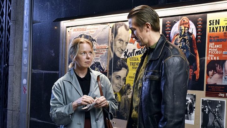 Filmstill from FALLEN LEAVES: A woman in a trench coat and a man in a leather jacket stand next to each other in front of film posters.