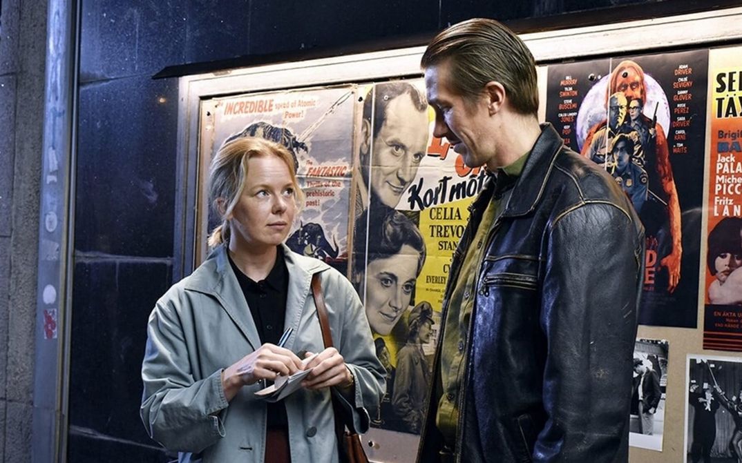 Filmstill from FALLEN LEAVES: A woman in a trench coat and a man in a leather jacket stand next to each other in front of film posters.