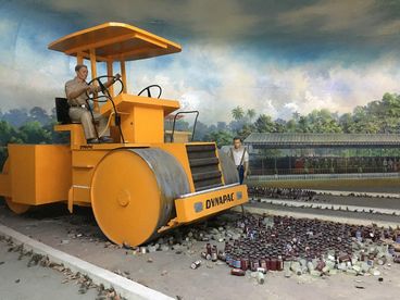 A behind-the-scenes shot. A diorama of two people, one of them riding a yellow tractor, seemingly driving over empty medicine bottles. 