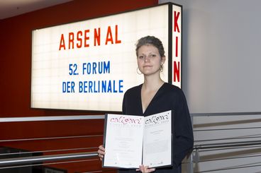Eine Frau steht vor einer Kino-Leuchttafel. Auf der Tafel steht „52. Berlinale Forum“. Sie hält eine Urkunde in die Kamera.