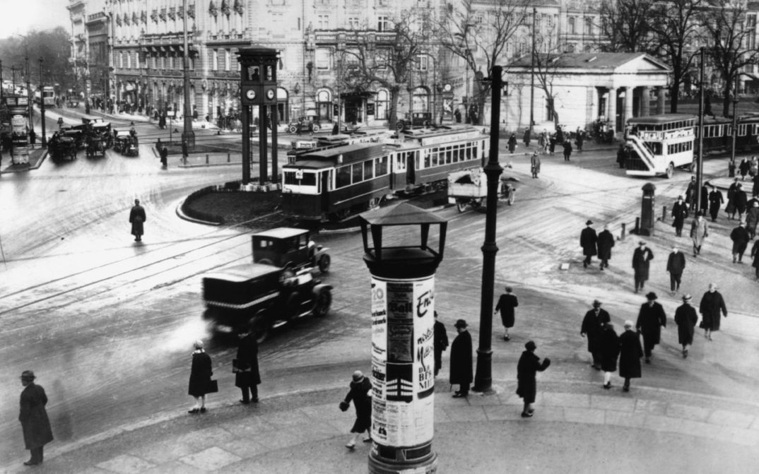 Eine Straßenkreuzung von oben; viele Passant*innen, eine Straßenbahn, Häuser und Autos.