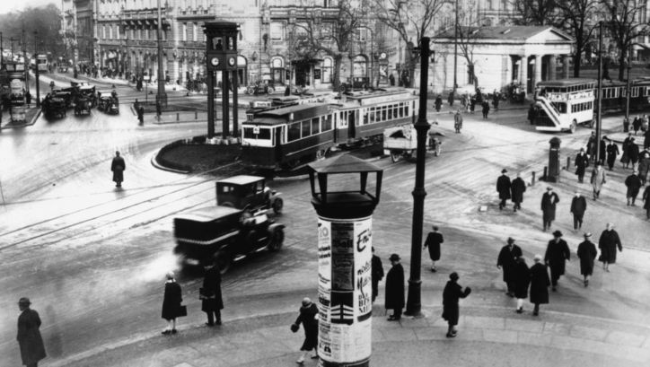 Eine Straßenkreuzung von oben; viele Passant*innen, eine Straßenbahn, Häuser und Autos.
