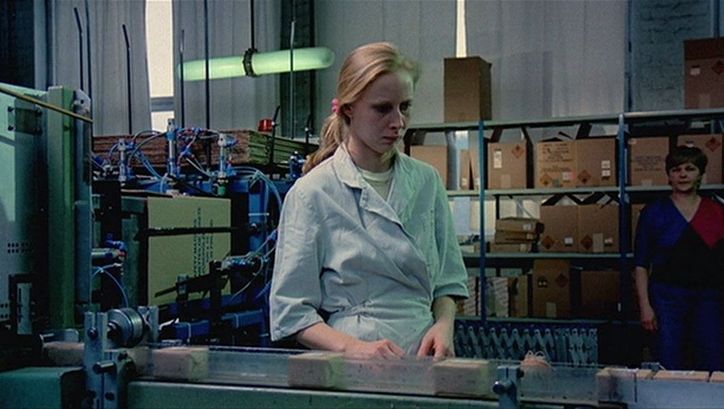 Film still from THE MATCH FACTORY GIRL: A young, blonde woman stands on the assembly line of a factory.