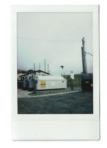 Polaroid of a power box outside of house, next to a dumpster and a parking lot.
