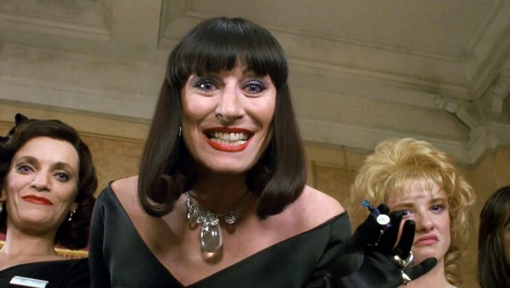  Film still from THE WITCHES: A woman wearing heavy makeup looks down at the floor with a menacing smile. Behind her are two other women.
