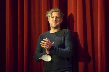 A man is standing in front of a red curtain, clapping.