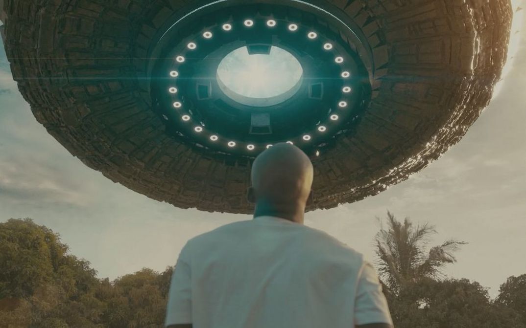 Person steht im Freien und blickt von hinten zu einem großen, schwebenden, kreisförmigen Objekt am Himmel.