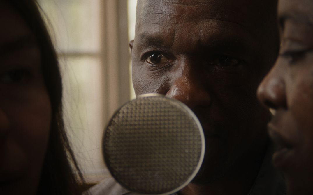 Film still from Robin Vanbesien’s film “hold on to her”. Two people lean towards a microphone that stands between them. A blurry unlit face can be seen on the left side of the frame.
