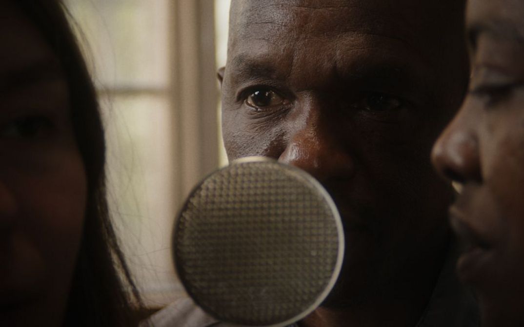 Film still from Robin Vanbesien’s film “hold on to her”. Two people lean towards a microphone that stands between them. A blurry unlit face can be seen on the left side of the frame.