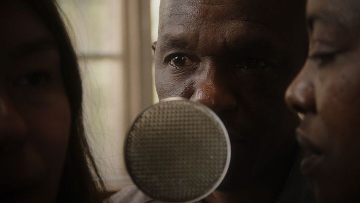 Filmstill aus dem Film „hold on to her“ von Robin Vanbesien. Zwei Personen beugen sich zu einem Mikrofon, das zwischen ihnen steht. Auf der linken Seite des Bildes ist ein verschwommenes, unbeleuchtetes Gesicht zu sehen.