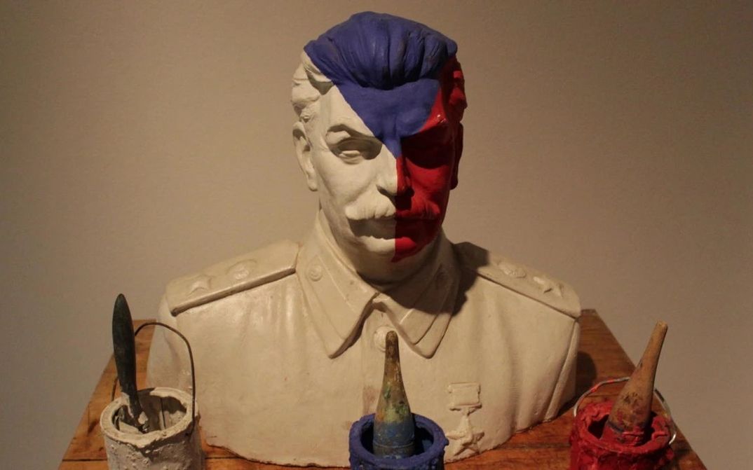 Filmstill aus THE DEATH OF STALINISM IN BOHEMIA: Eine Büste Stalins ist in den Farben der tschechischen Flagge bemalt – weiß, blau, rot. Auf dem Tisch vor der Büste stehen die drei Farbeimer mit Pinseln.