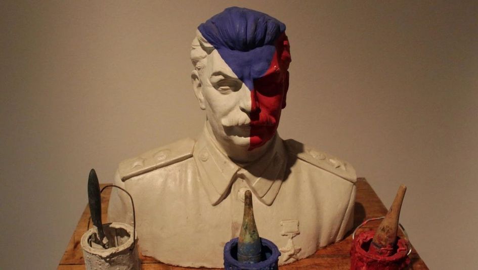 Film still from THE DEATH OF STALINISM IN BOHEMIA: A bust of Stalin is painted in the colors of the Czech flag—white, blue, and red. On the table in front of the bust are three paint buckets with brushes.