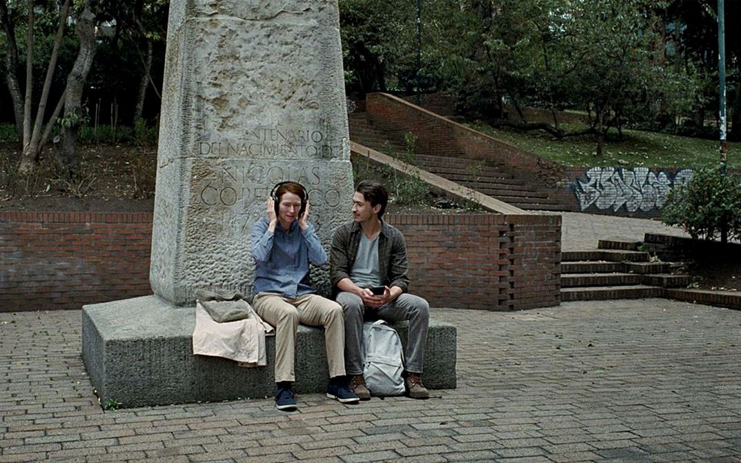 Filmstill aus MEMORIA: Eine Frau und ein Mann sitzen auf dem Sockel eines angeschnittenen Denkmals. Die Frau hat Kopfhörer aufgesetzt, der Mann schaut sie an.