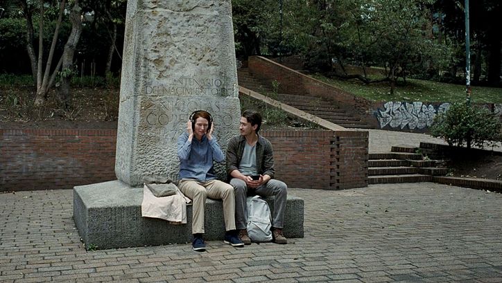 Filmstill aus MEMORIA: Eine Frau und ein Mann sitzen auf dem Sockel eines angeschnittenen Denkmals. Die Frau hat Kopfhörer aufgesetzt, der Mann schaut sie an.