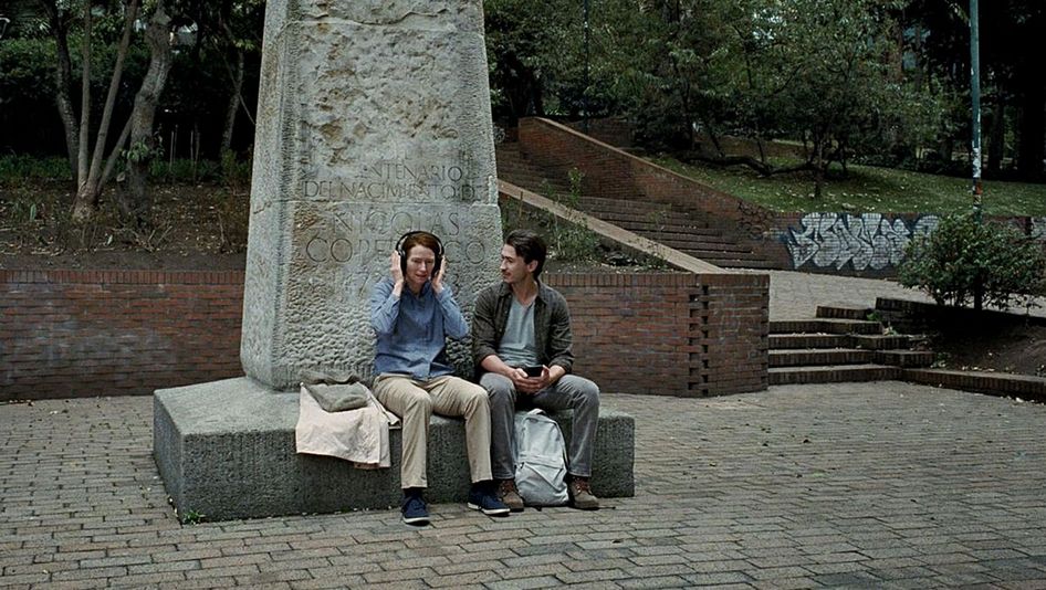 Filmstill aus MEMORIA: Eine Frau und ein Mann sitzen auf dem Sockel eines angeschnittenen Denkmals. Die Frau hat Kopfhörer aufgesetzt, der Mann schaut sie an.