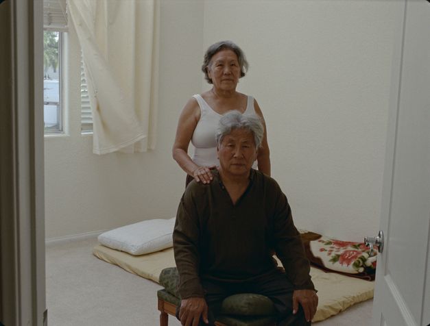 Film still from Tenzin Phuntsog’s "Pala Amala". An elderly couple in the middle of a room in front of a mattress.