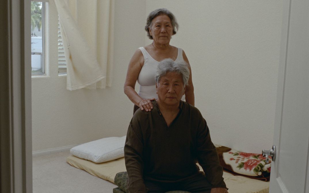 Film still from Tenzin Phuntsog’s "Pala Amala". An elderly couple in the middle of a room in front of a mattress.