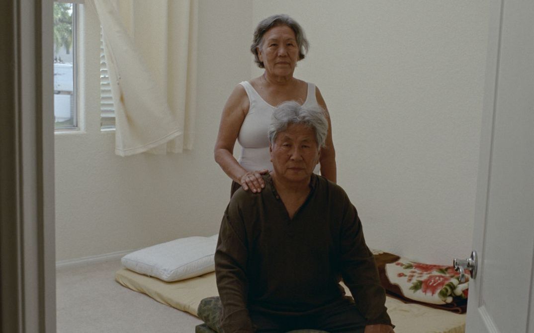 Film still from Tenzin Phuntsog’s "Pala Amala". An elderly couple in the middle of a room in front of a mattress.