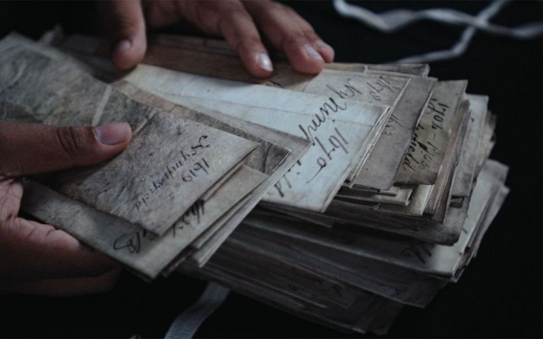 Filmstill aus dem Film „Black Strangers“ von Dan Guthrie. Zwei Hände streifen durch einen Stapel vergilbter Akten.