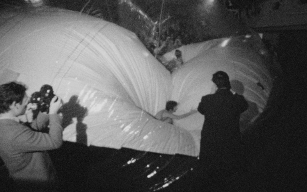 Filmstill aus EXPRMNTL 4 KNOKKE: Schwarzweiß Bild in einem dunklen Innenraum: Zwei Personen filmen mit Kameras, während mehrere andere auf großen, glänzenden, aufgeblasenen Ballonformen stehen oder darauf balancieren, die den Raum fast vollständig ausfüllen.