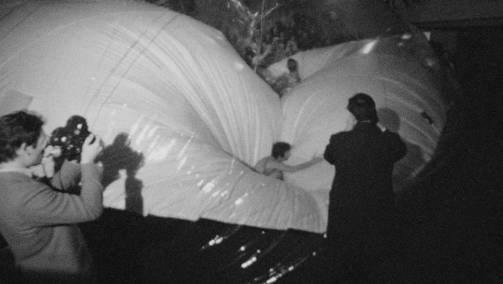 Filmstill aus EXPRMNTL 4 KNOKKE: Schwarzweiß Bild in einem dunklen Innenraum: Zwei Personen filmen mit Kameras, während mehrere andere auf großen, glänzenden, aufgeblasenen Ballonformen stehen oder darauf balancieren, die den Raum fast vollständig ausfüllen.