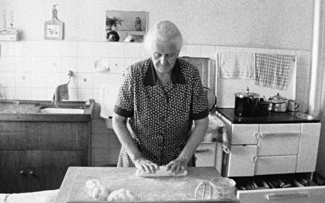 Filmstill aus FAMILIENGRUFT: Eine alte Frau steht in einer einfachen Küche und knetet einen Brotteig.