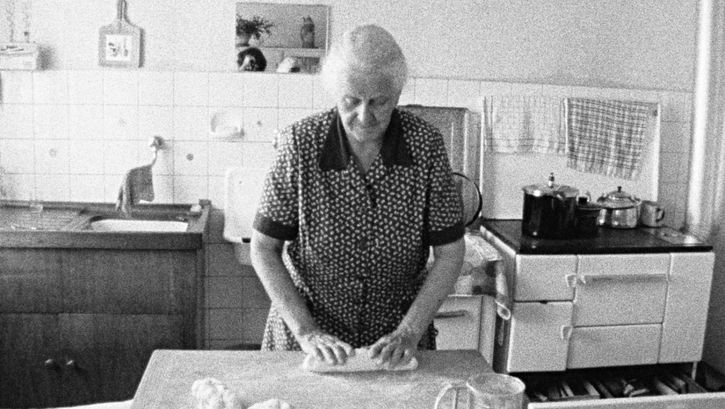 Filmstill aus FAMILIENGRUFT: Eine alte Frau steht in einer einfachen Küche und knetet einen Brotteig.