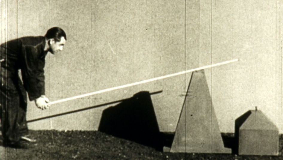 Filmstill aus MONGOLOID: Ein Mann eine Stange über ein sich am Boden befindendes Objekt.