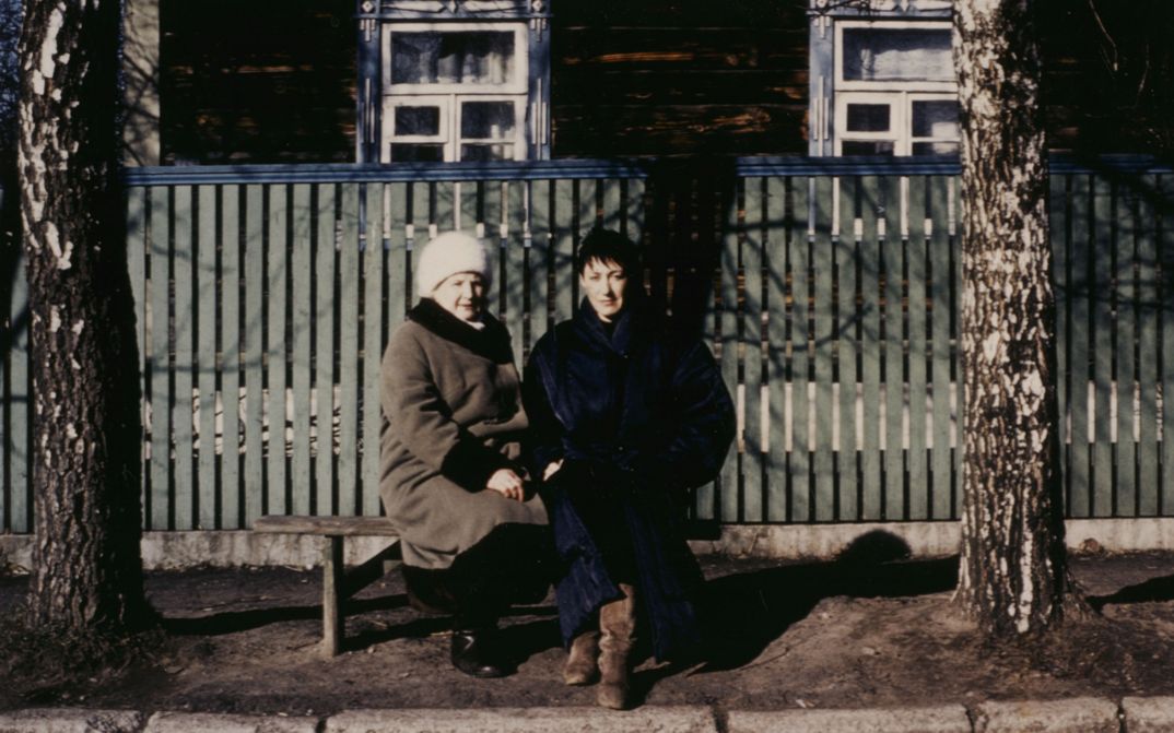 Filmstill aus BEFREIER UND BEFREITE. Zwei Frauen sitzen auf einer Bank an einer Straße und blicken in die Kamera.