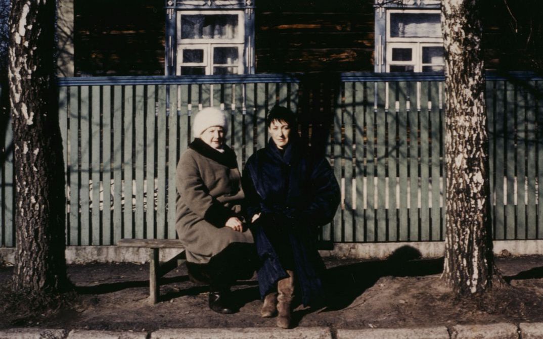 Filmstill aus BEFREIER UND BEFREITE. Zwei Frauen sitzen auf einer Bank an einer Straße und blicken in die Kamera.