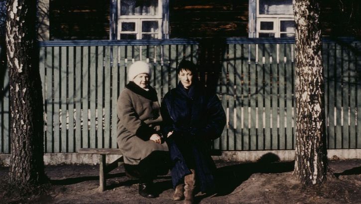 Filmstill aus BEFREIER UND BEFREITE. Zwei Frauen sitzen auf einer Bank an einer Straße und blicken in die Kamera.