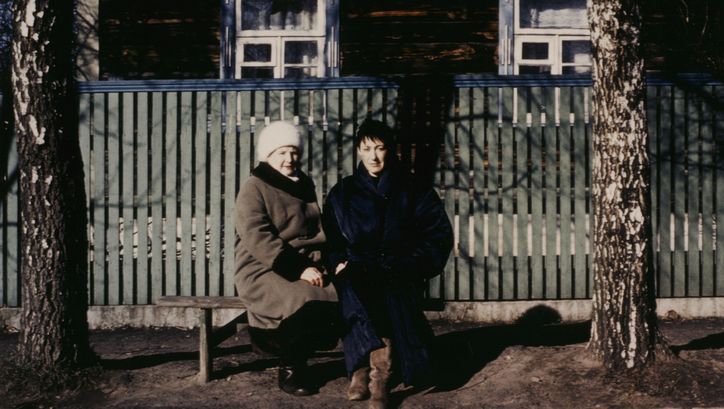 Filmstill aus BEFREIER UND BEFREITE. Zwei Frauen sitzen auf einer Bank an einer Straße und blicken in die Kamera.