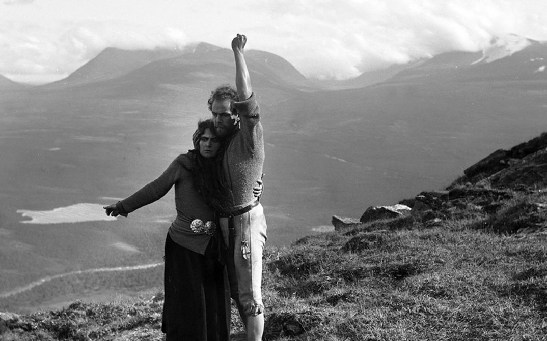 Filmstill aus BERG-EYVIND UND SEINE FRAU: Ein Mann und eine Frau stehen in einer einsamen Gebirgslandschaft. Sie halten einander fest und haben je einen Arm von sich gestreckt. 