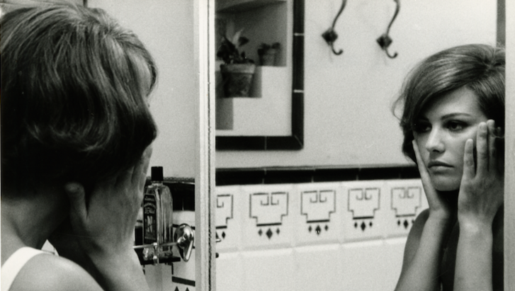 Film still from GLI INDIFFERENTI: A woman looks at herself in the bathroom mirror and holds her hands to her cheeks.