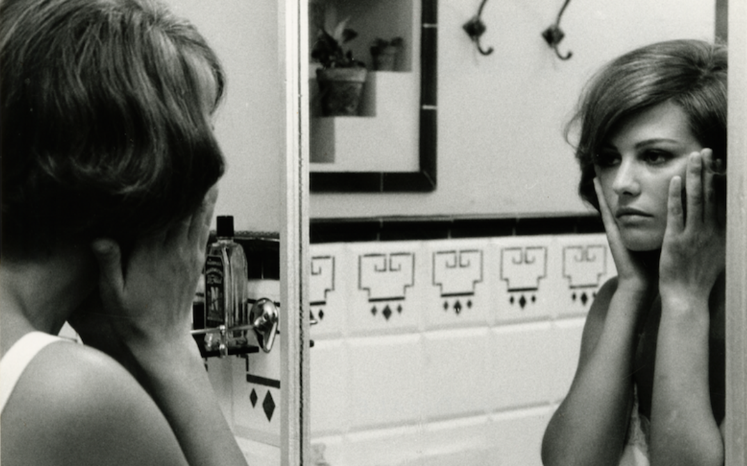 Film still from GLI INDIFFERENTI: A woman looks at herself in the bathroom mirror and holds her hands to her cheeks.