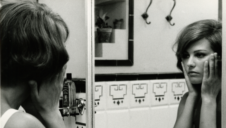 Filmstill aus GLI INDIFFERENTI: Eine Frau betrachtet sich im Badezimmerspiegel und hält die Hände an die Wangen.